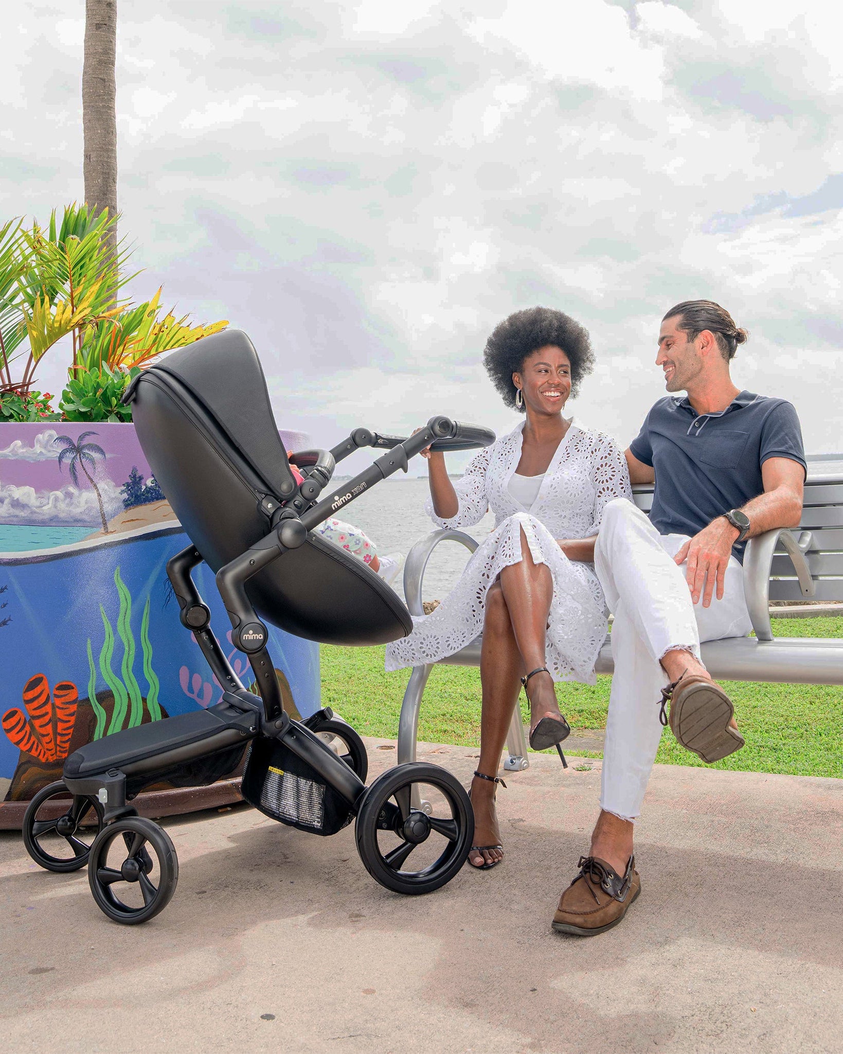 Couple sitting on a bench with a black xari stroller in a park setting.