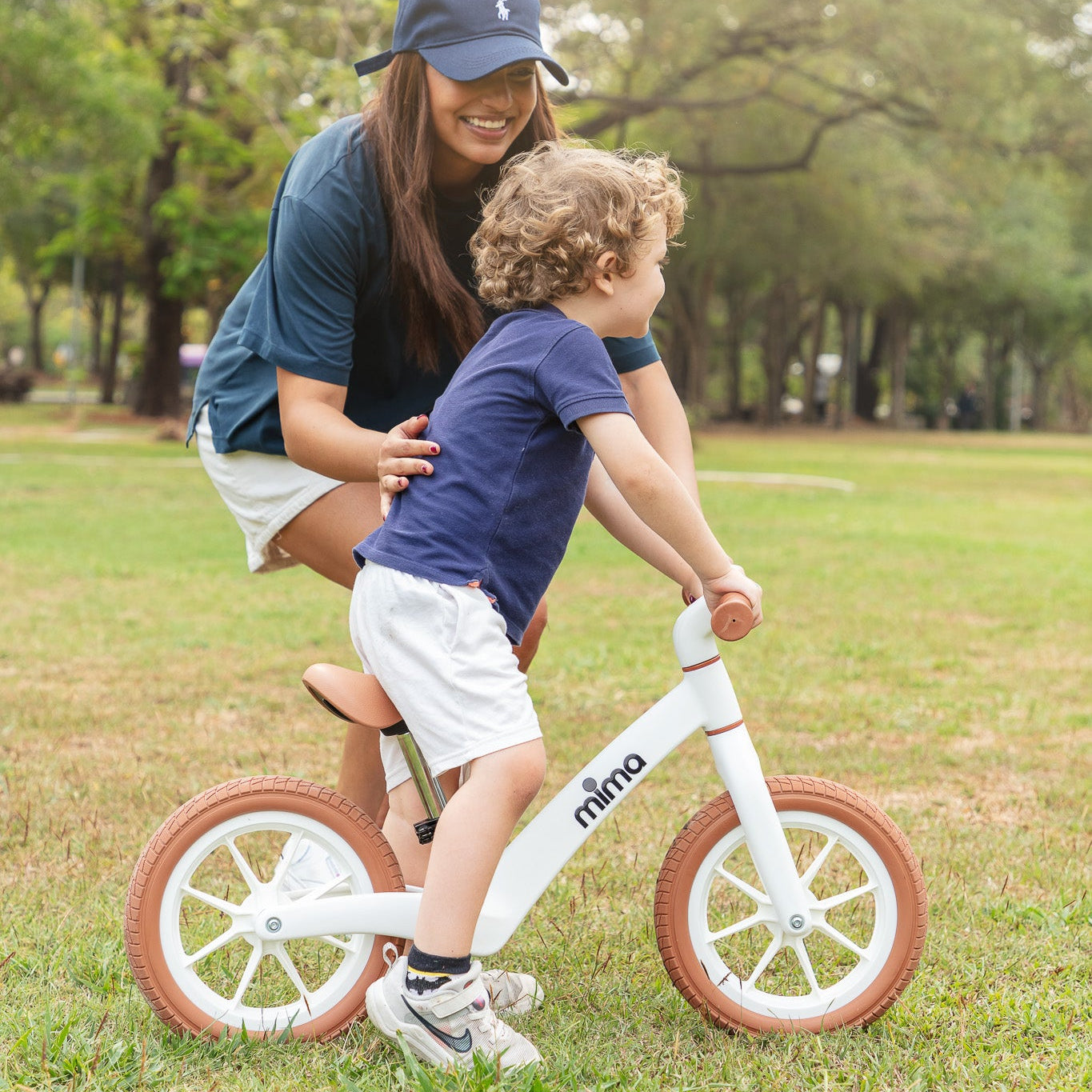 A smiling mother in a cap guiding her toddler riding a white mima zoom lite balance bike on a grass field.