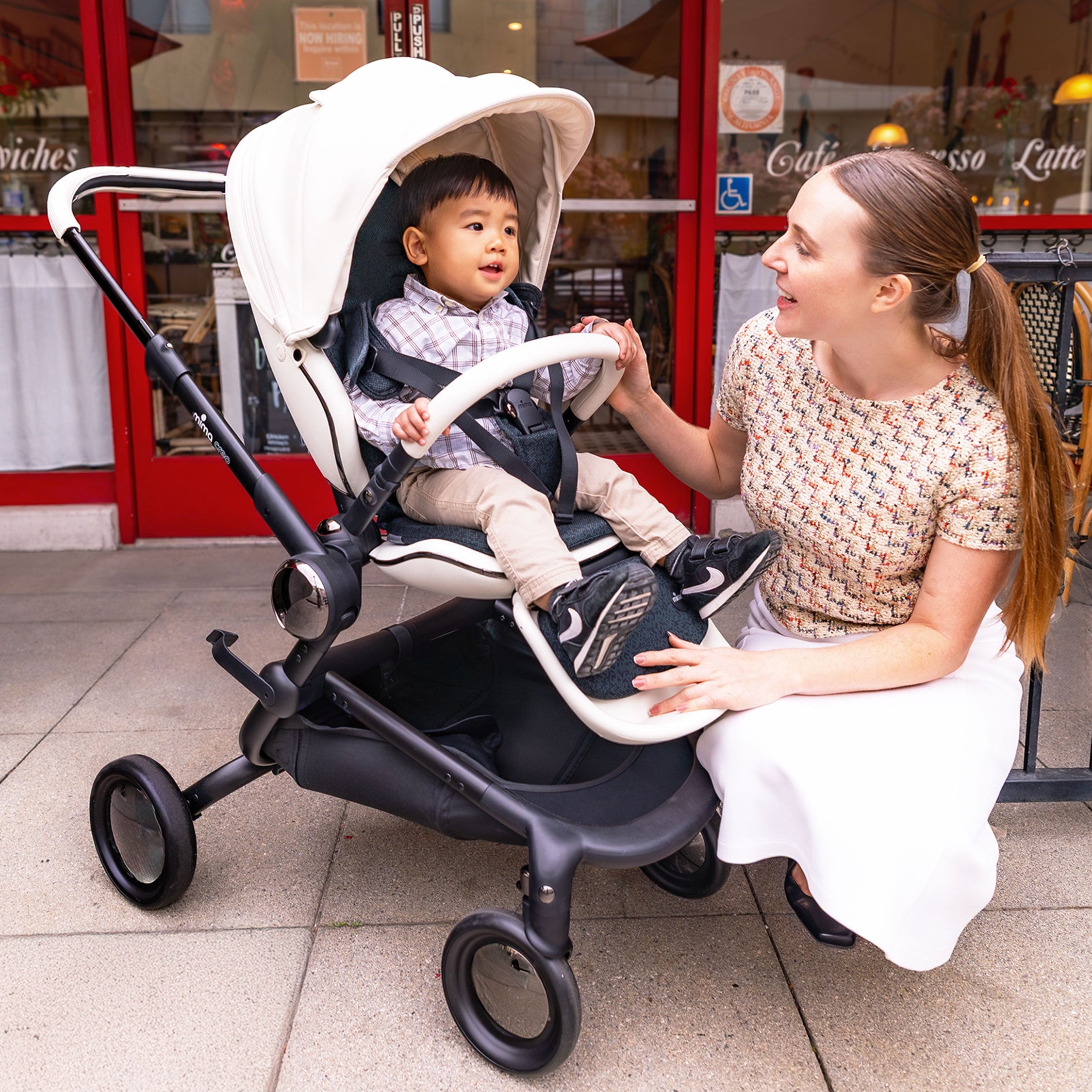 Woman sitting on a sidewalk with a child in a  creo white stroller, with a storefront in the background.