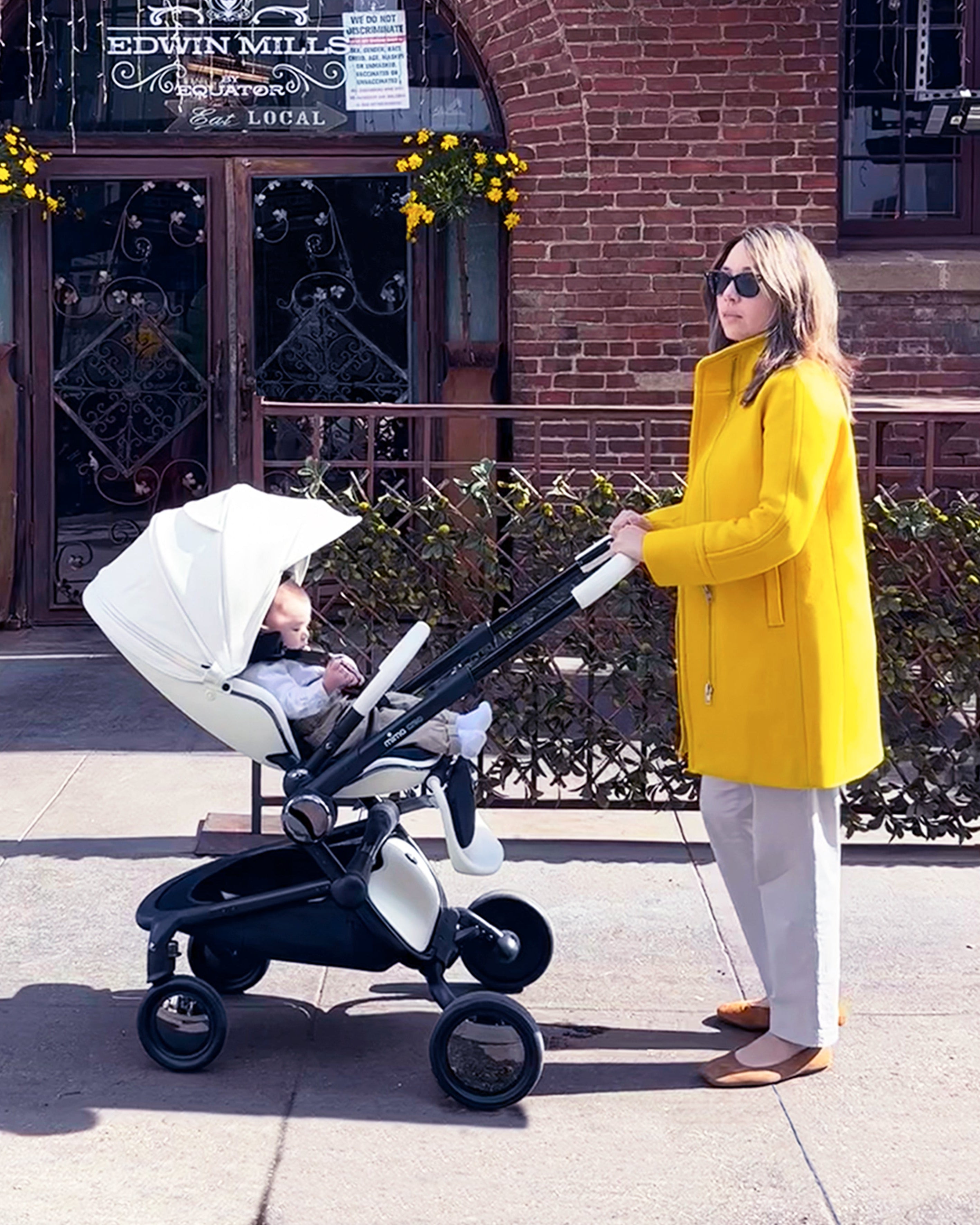 caregiver in yellow coat standing with mima creo compact stroller in white on a city street