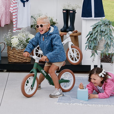 Two young children smiling and playing outdoors with white and green mima zoom lite balance bikes.